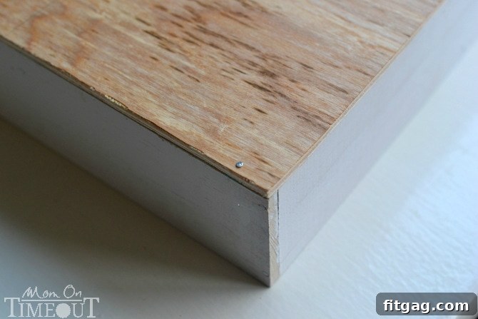 A close-up view of a hand using a hammer to nail the plywood base to the white wooden dividers of the DIY drawer organizer, ensuring a sturdy construction.