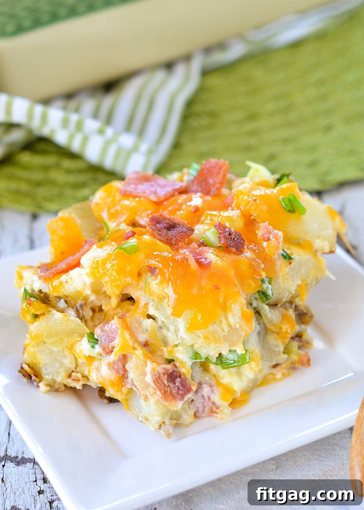 A generous serving of Twice Baked Potato Casserole on a white square plate, with the casserole dish visible in the background. The casserole is topped with melted cheese, crispy bacon, and chopped green onions.