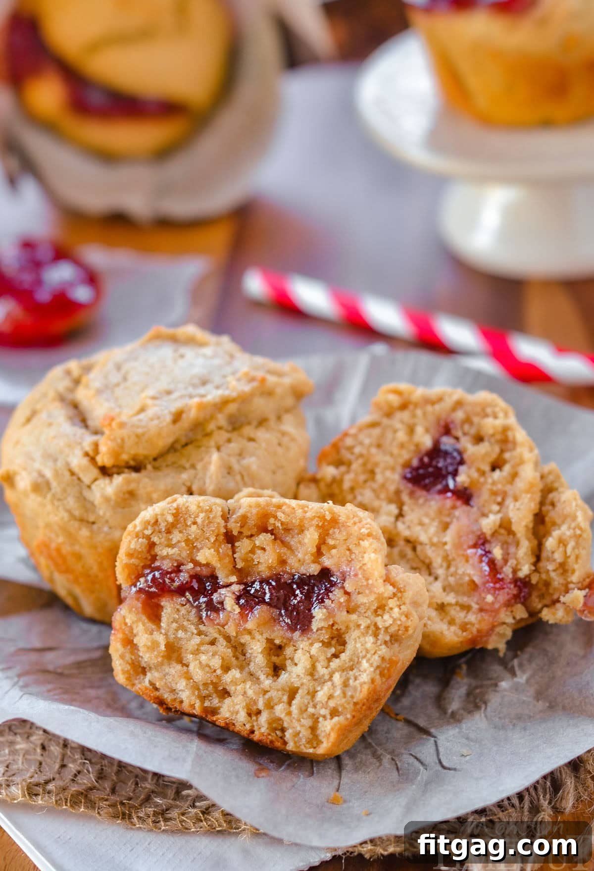 Two peanut butter and jelly muffins on a piece of parchment. One muffin has been split in half to reveal the jelly center.