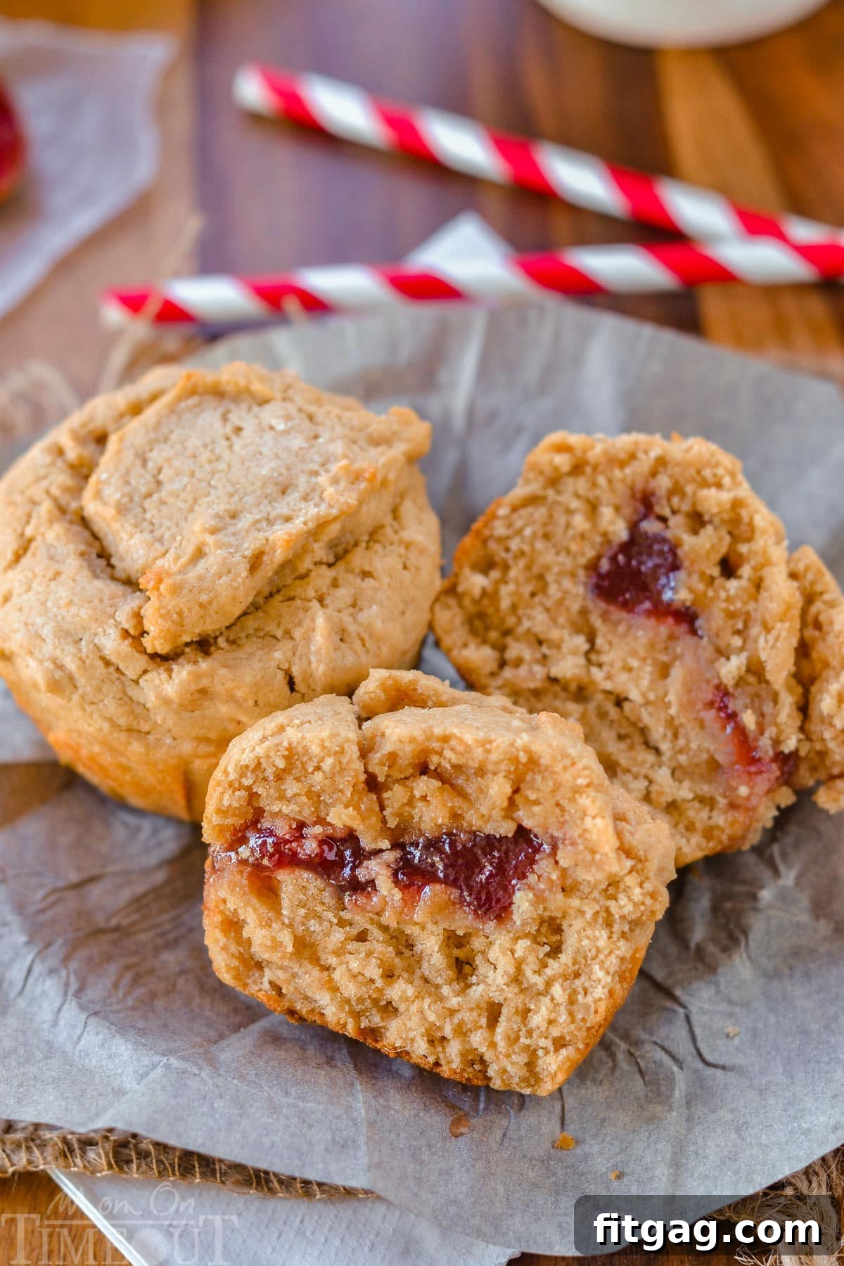 Two peanut butter and jelly muffins on parchment paper, one sliced open to reveal the vibrant jelly filling.