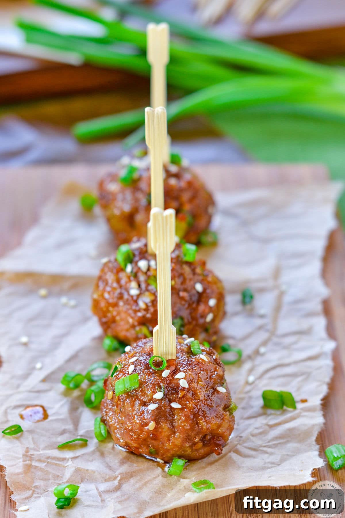 Glazed Teriyaki Meatballs 2 Three teriyaki meatballs lined up on parchment paper, topped with green onions and sesame seeds. Toothpicks have been inserted into the top of each meatball.