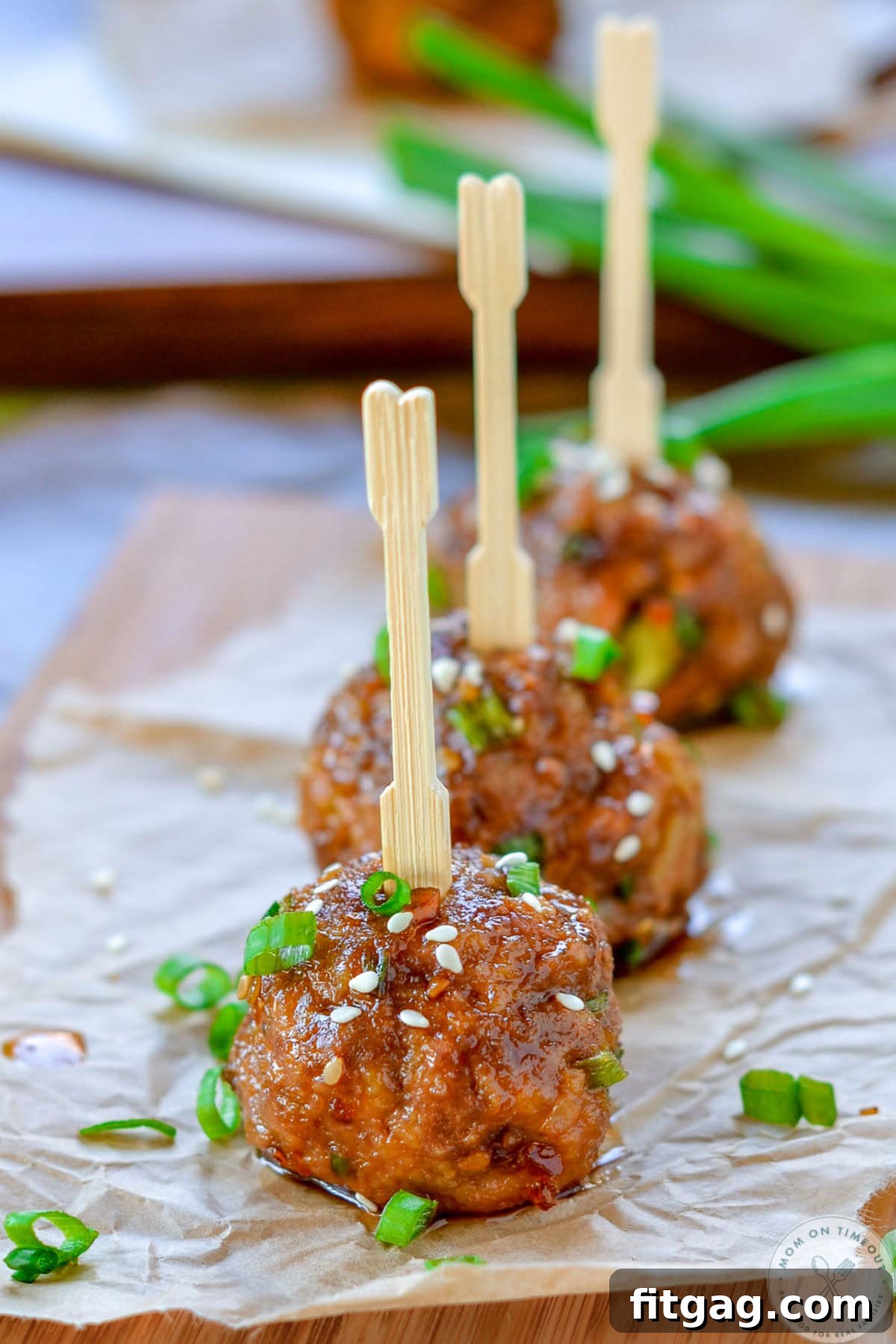 Glazed Teriyaki Meatballs 5 Three teriyaki meatballs lined up on parchment paper, topped with green onions and sesame seeds. Toothpicks have been inserted into the top of each meatball.