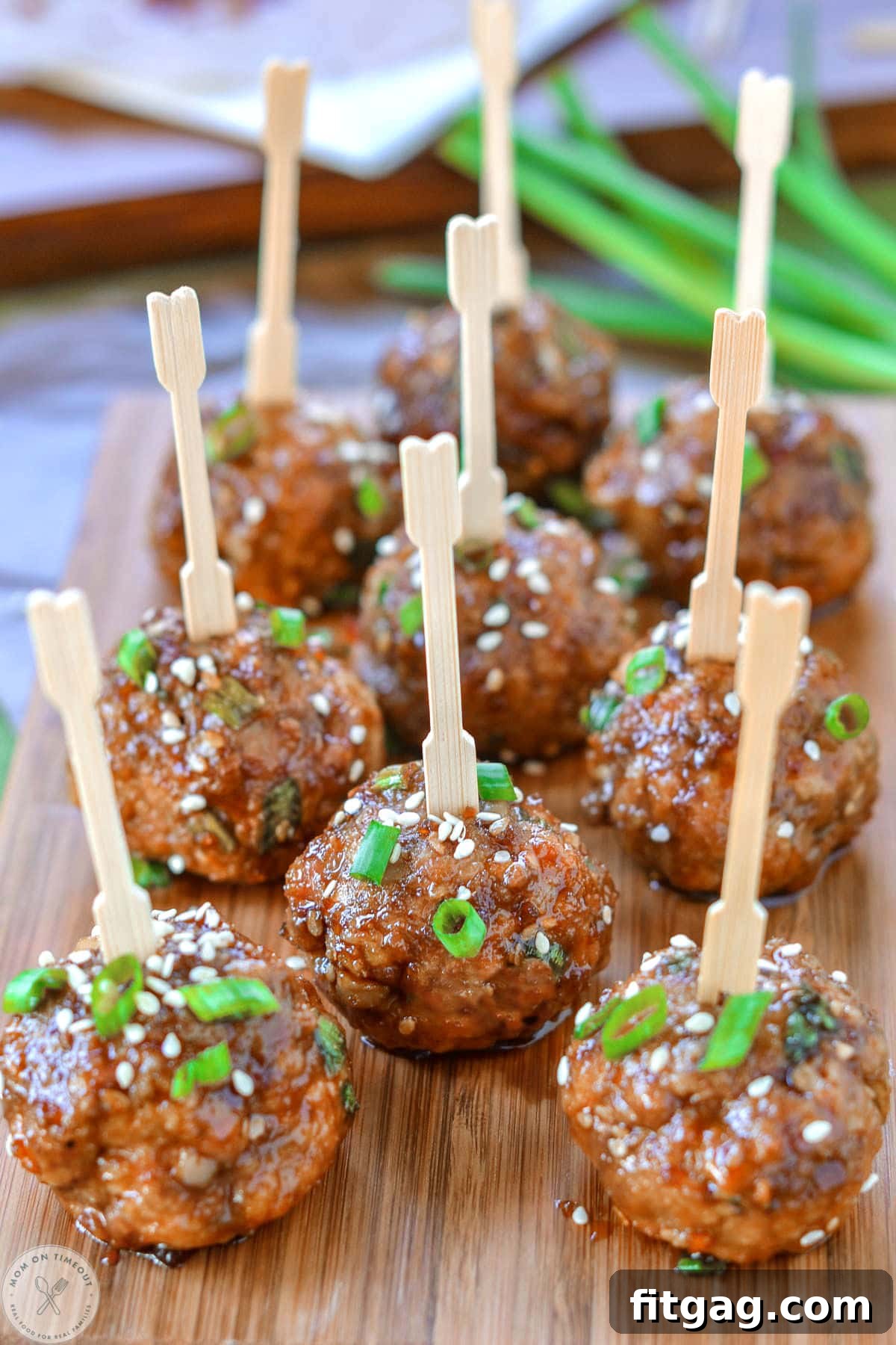Glazed Teriyaki Meatballs 6 Nine teriyaki meatballs on wood cutting board topped with green onions and sesame seeds. Each meatball has a toothpick in it.