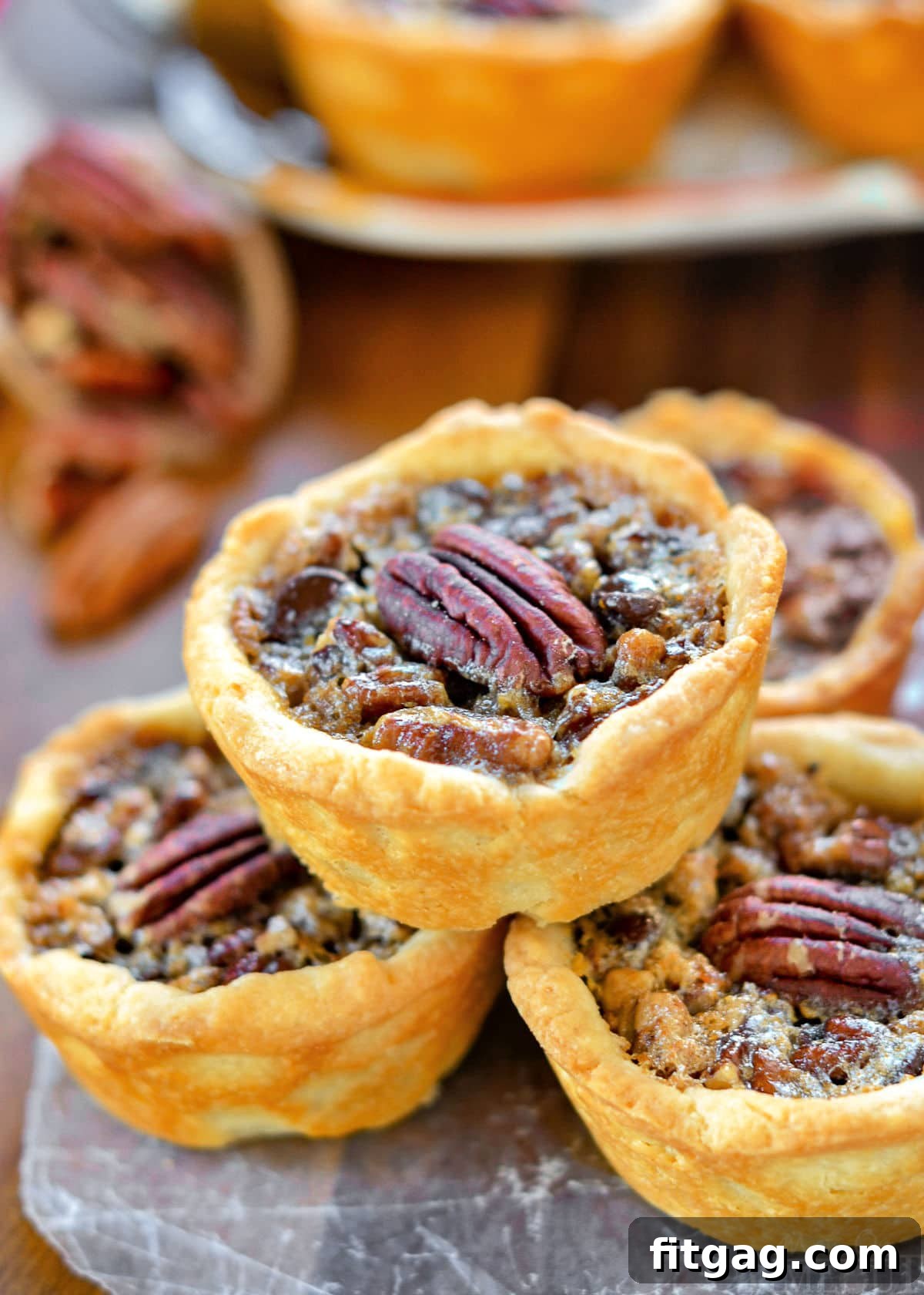 Pecan Pie Bites 2 Four golden-brown mini pecan pies stacked elegantly, topped with glistening pecan halves, against a blurred background of more mini pies. This image perfectly captures the festive and delicious nature of the holiday dessert.