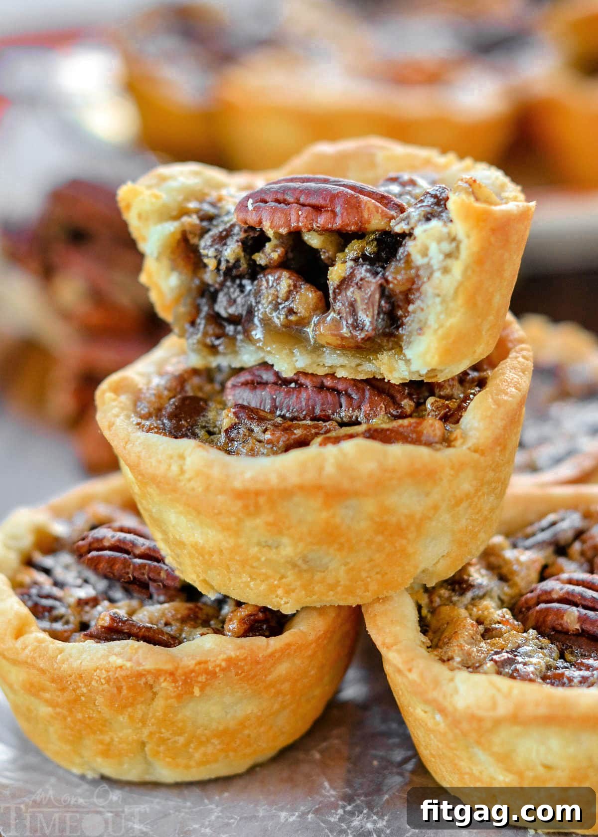 Pecan Pie Bites 7 Five beautifully baked mini pecan pies, stacked in a pyramid shape, with more pies in the background. The top pie is cut in half, revealing the rich, dense filling and crunchy pecans. Each pie is adorned with a perfect pecan half on top.