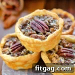 Four mini pecan pies stacked in a pyramid with more pies in the background. each mini pie is stopped with a pecan half.