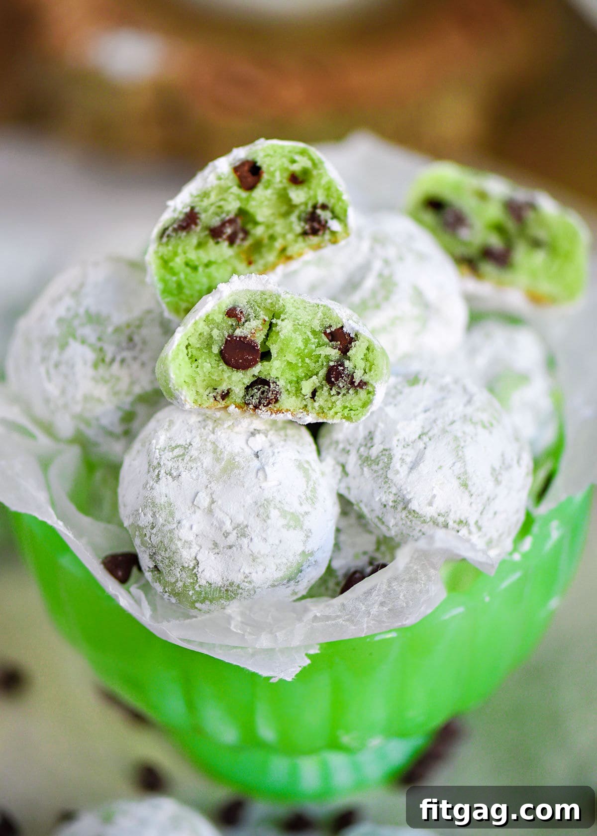 mint chocolate chip snowball cookies in a small green glass pedestal bowl lined with wax paper. A few cookies are broken in half to show the green interior and mini chocolate chips.