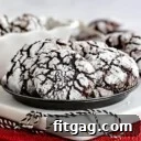 plate of chocolate crinkle cookies sitting on white wood board on red sparkly placemat. black and white twine sitting on board and more cookies can be seen in the background.