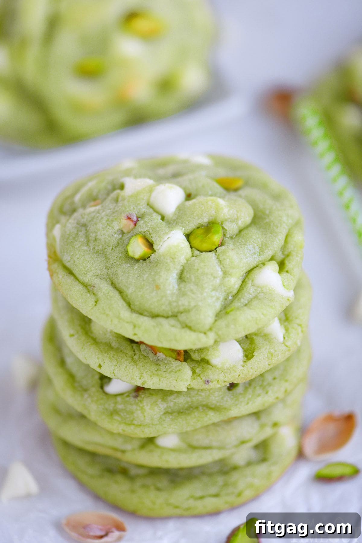 Velvety Pistachio Pudding Cookies 2 A stack of five vibrant green pistachio cookies adorned with white chocolate chips and chopped pistachios on white parchment paper.
