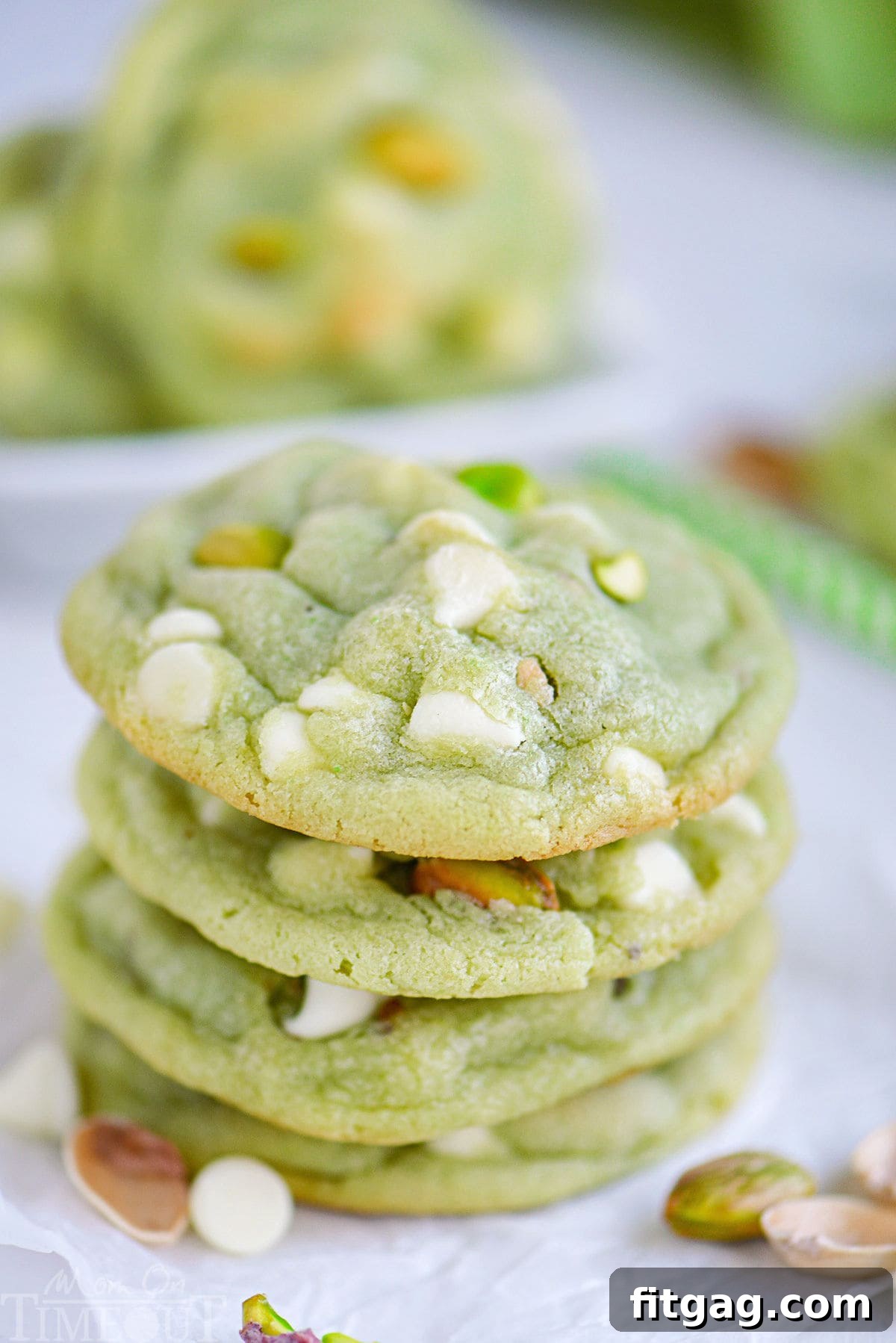 Velvety Pistachio Pudding Cookies 5 A stack of four soft pistachio pudding cookies on white parchment paper, featuring prominent white chocolate chips and chopped pistachios.