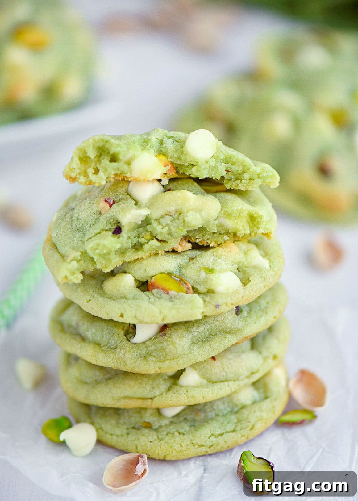Velvety Pistachio Pudding Cookies 6 A stack of pistachio pudding cookies with one broken in half to reveal the soft, chewy interior, white chocolate chips, and pistachios. More cookies are blurred in the background.