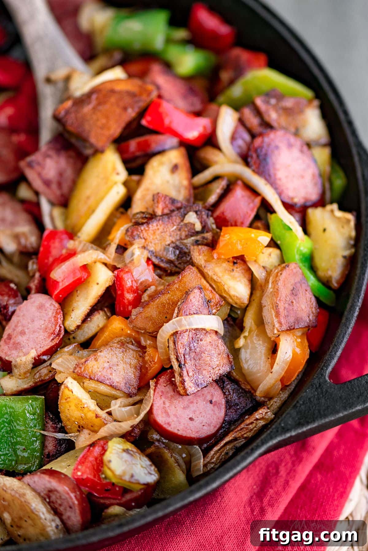 Kielbasa and potatoes in a cast iron skillet ready to be enjoyed. Skillet is resting on a red napkin. Peppers, onions, potatoes and kielbasa are all cut into bite sized pieces.