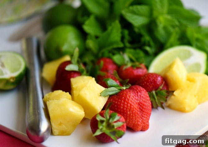 A vibrant Pineapple Strawberry Mojito being prepared in a glass, with muddled strawberries, pineapple, mint, and lime at the bottom, ready for the liquids to be added.