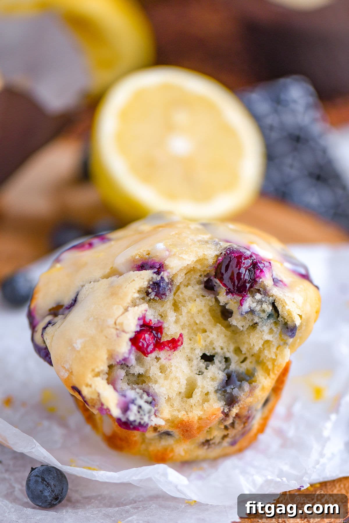 Cream Cheese Stuffed Blueberry Lemon Muffins 2 A perfectly baked Blueberry Lemon Cream Cheese Muffin with a bite taken out, revealing its moist interior packed with blueberries. It rests on white parchment paper, with a lemon half visible in the soft-focus background, suggesting freshness.