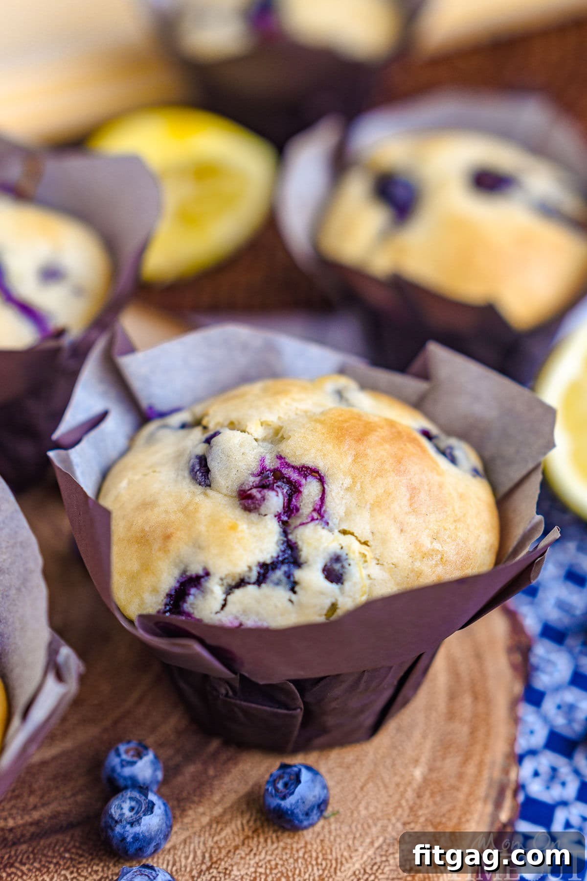 A single Blueberry Lemon Muffin in a tulip parchment liner rests on a rustic wood board, accompanied by a scattering of fresh blueberries.