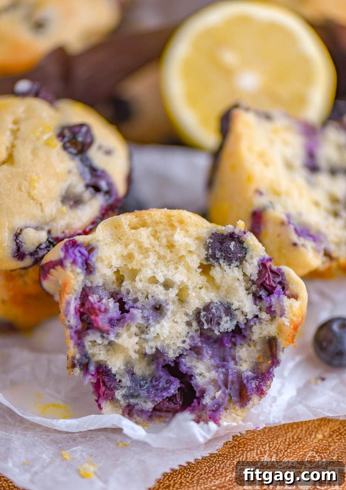 Cream Cheese Stuffed Blueberry Lemon Muffins 4 A Blueberry Lemon Cream Cheese Muffin, artfully split in half to reveal its tender, blueberry-studded interior, is displayed on white parchment paper. More perfectly baked muffins and fresh lemon halves are softly blurred in the background, creating an inviting scene.