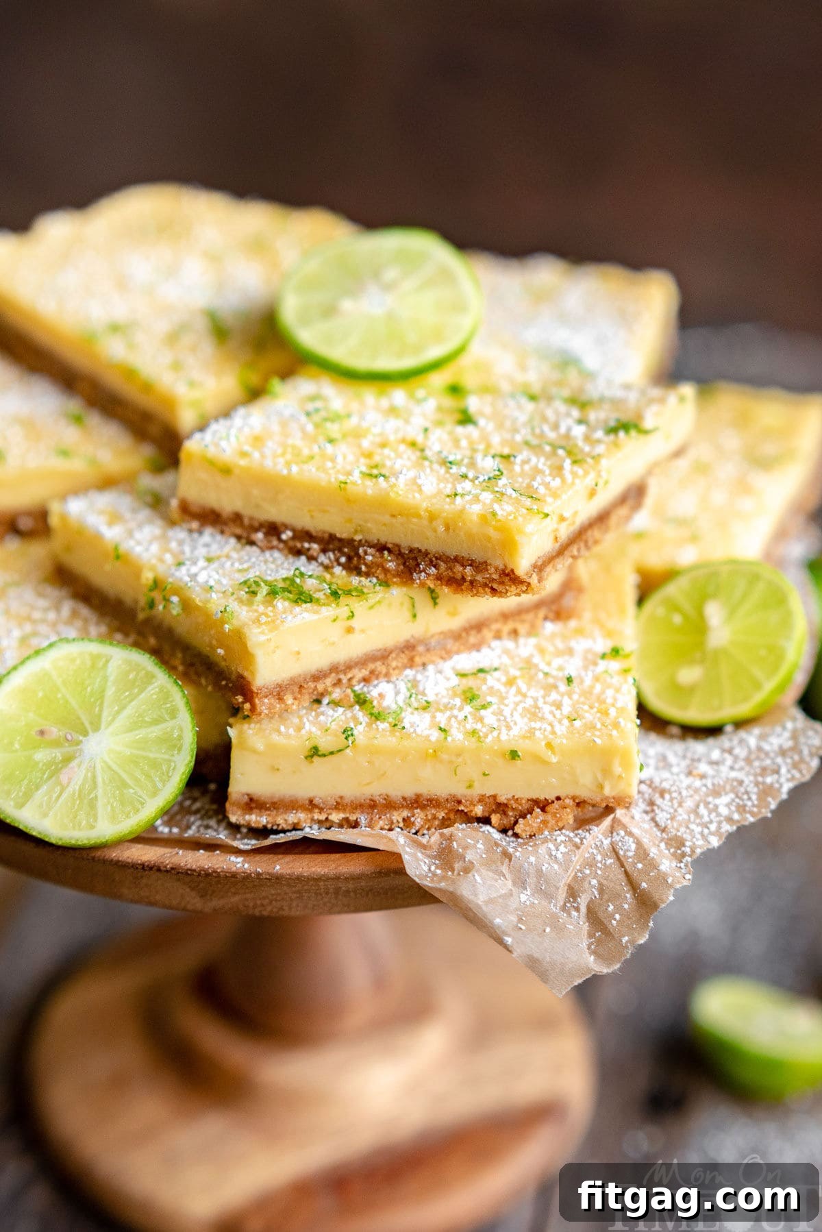 Tangy Key Lime Bars 2 A stunning wood cake stand overflowing with perfectly cut Key Lime Pie Bars, adorned with vibrant key lime zest and a light veil of powdered sugar. Fresh lime slices are artfully scattered around the base, enhancing the visual appeal.