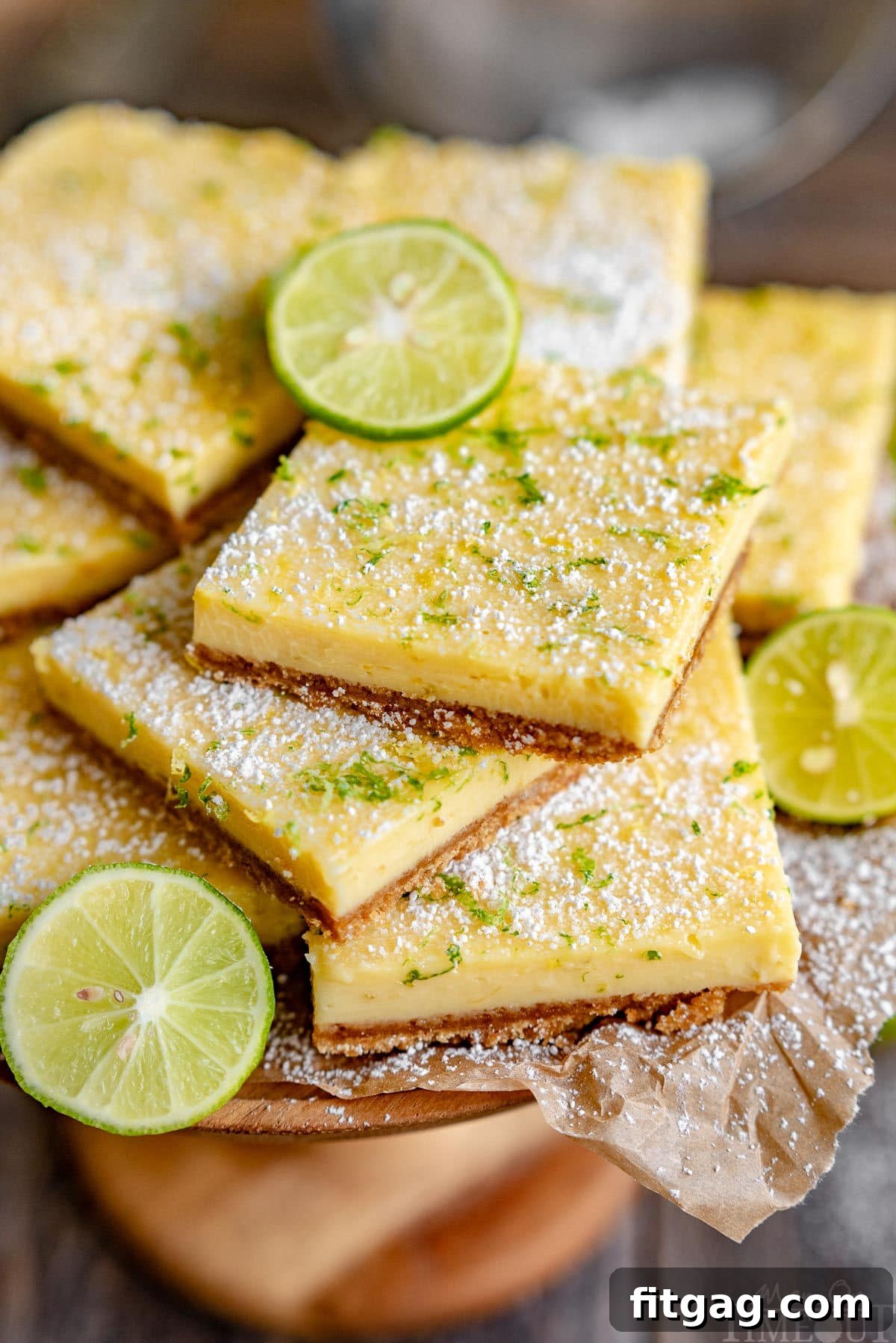 Tangy Key Lime Bars 6 A close-up shot of several Key Lime Pie Bars artfully arranged on a wooden cake stand, lined with parchment paper. The bars are exquisitely topped with a blend of lemon and lime zest, along with a fine dusting of powdered sugar.