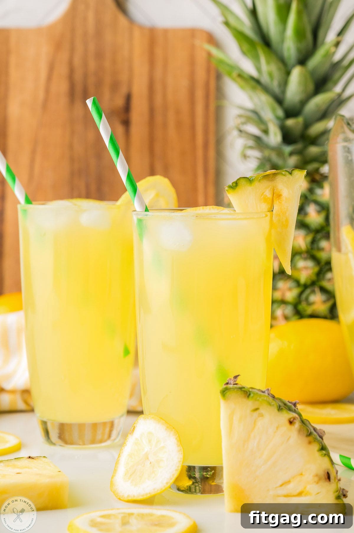 Two glasses filled with pineapple lemonade. Straws in glasses with fresh pineapple in the background.