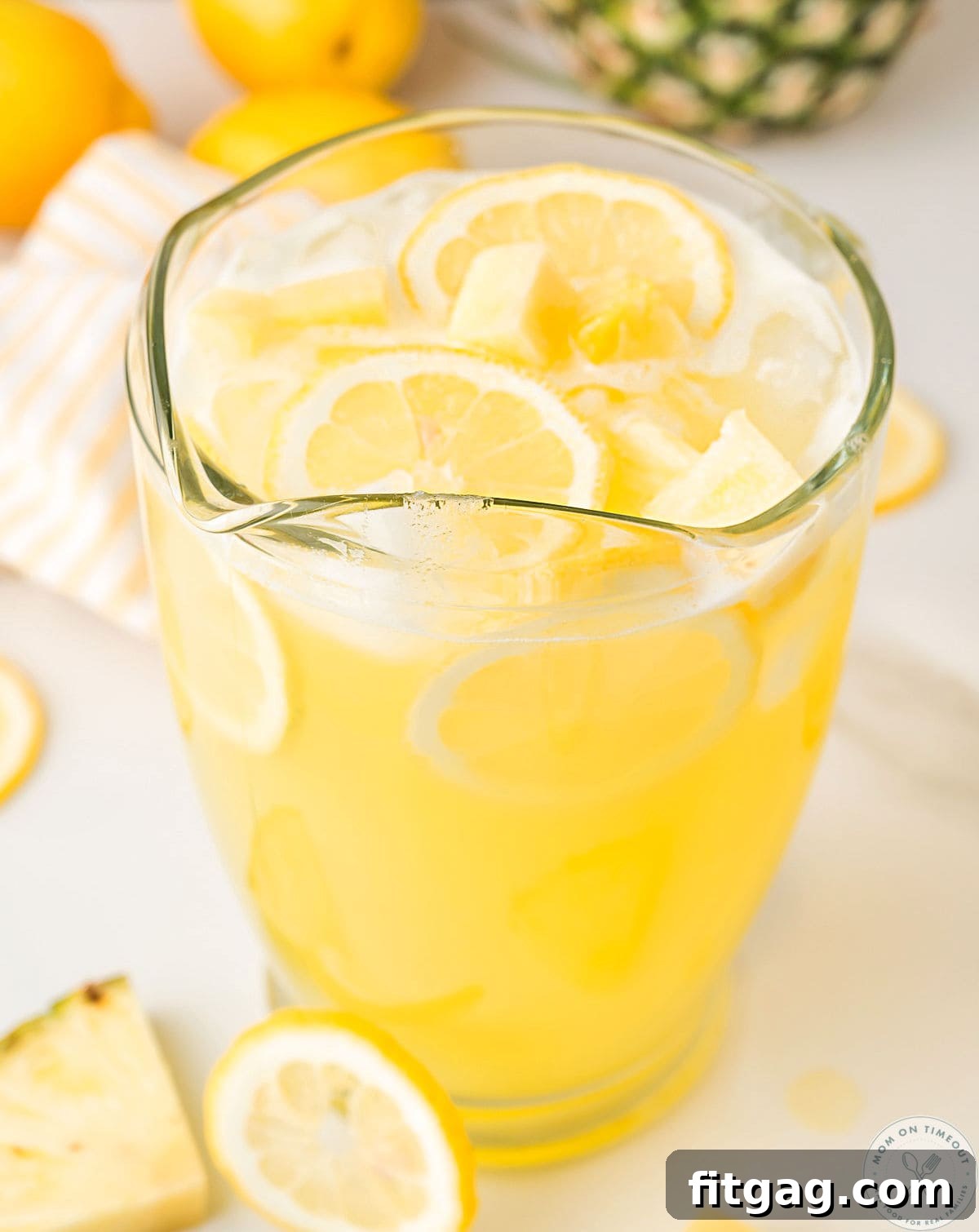 Angled overhead shot of pineapple lemonade recipe in glass pitcher garnished with lemon slices and pineapple chunks.